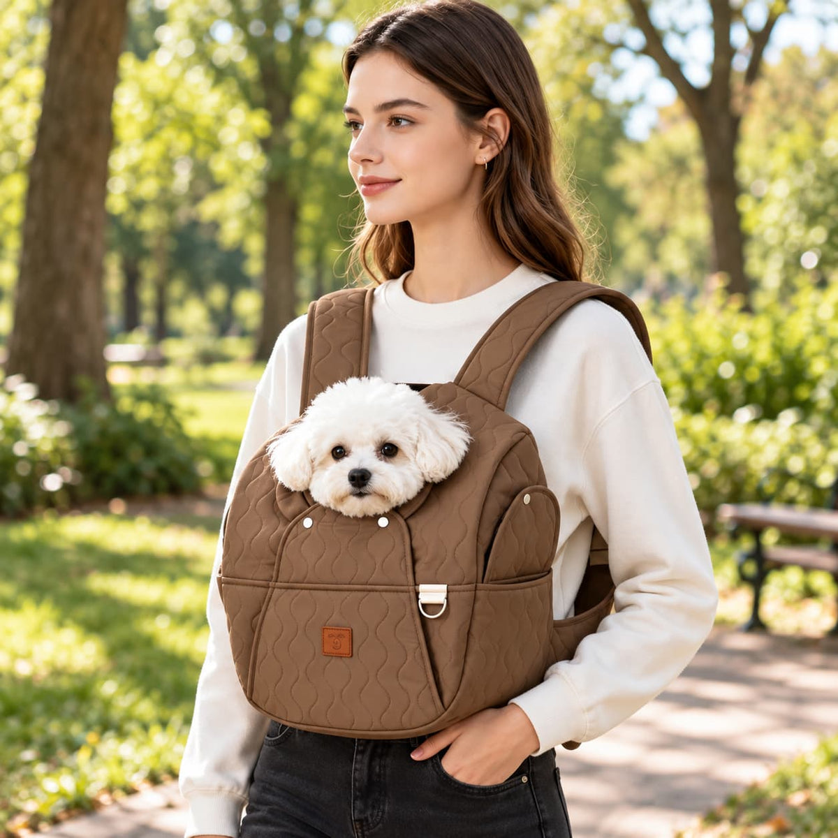 Woman carrying a small white dog in a brown front pet carrier while walking in a park