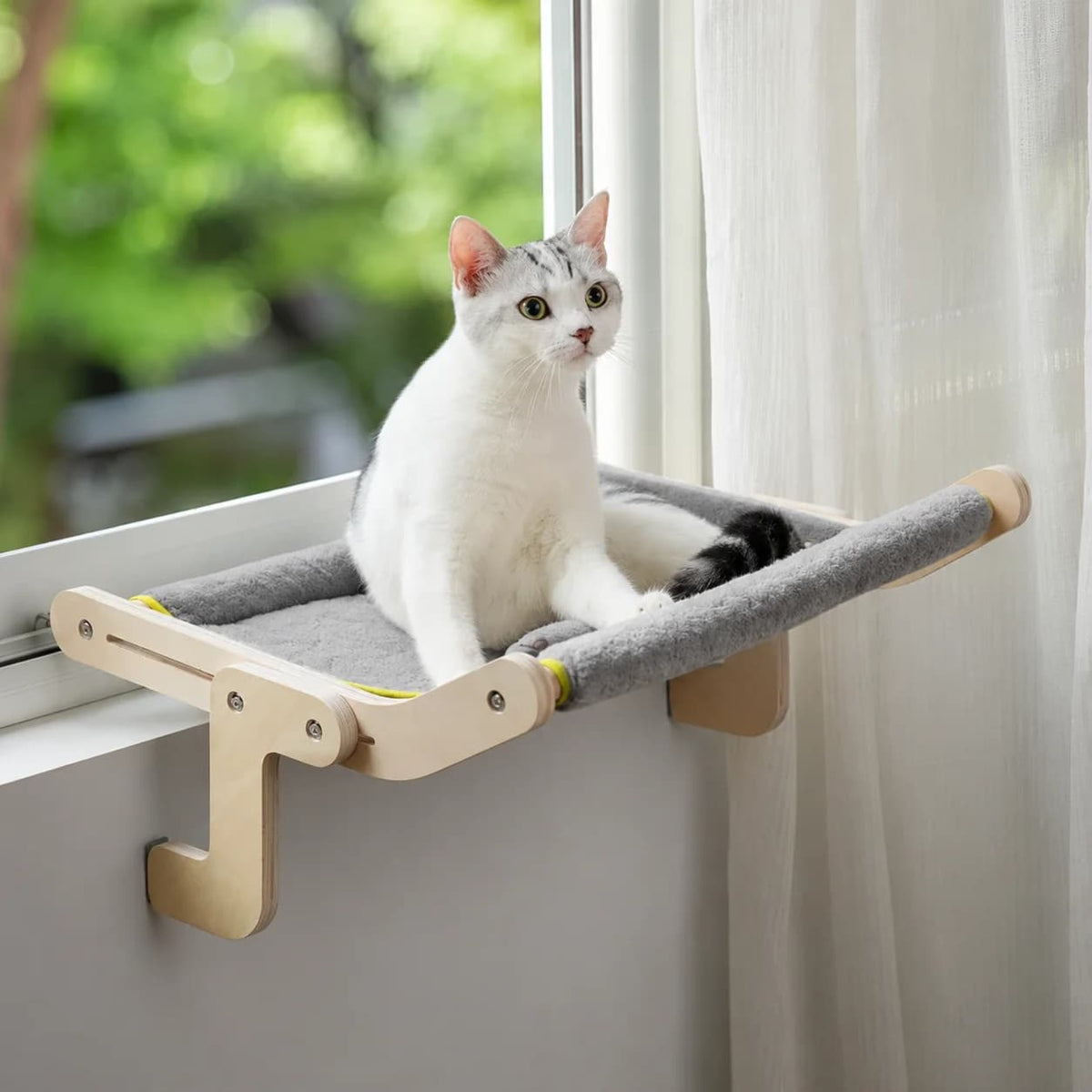 White cat sitting comfortably on a grey window mounted hammock bed with wooden supports.